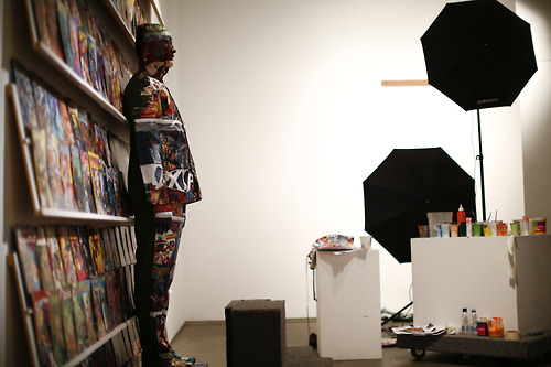 Liu Bolin, a Chinese artist with camouflage paint applied all over him, stands in front of a shelf lined with comic books as part of a series of performances in Caracas